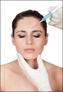 A woman with her eyes closed with two gloved hands preparing to inject a syringe into her facial skin.